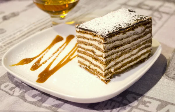 A piece of honey cake with custard and chocolate honey shortcakes on decorated plate and glass of brandy on background.