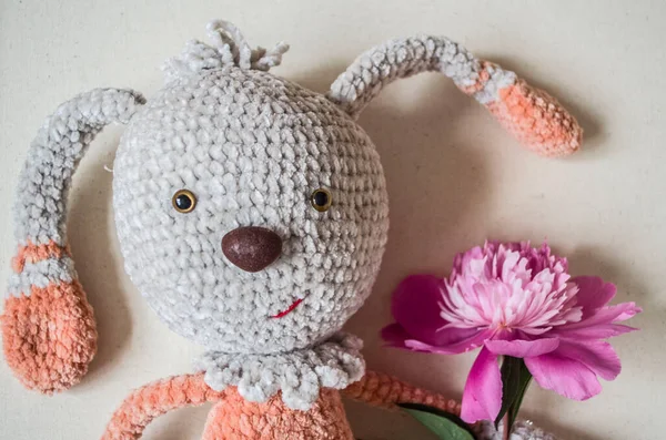 Crocheted hare with pink peony flower on wooden background. Handmade toy
