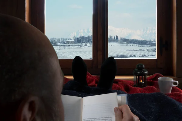 a man reads a book in front of the window on a winter day