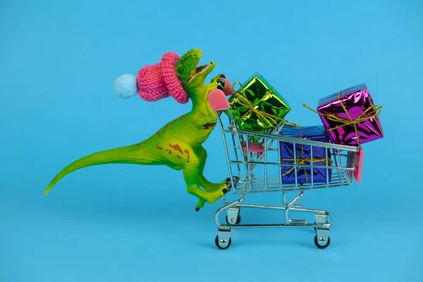 Komik plastik oyuncak dinozor küçük örgü şapkalı ve hediye kutularıyla dolu mavi arka planda alışveriş konseptini kullanıyor.