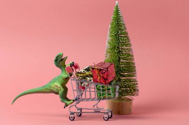 Komik plastik oyuncak dinozor hediye kutularıyla dolu alışveriş arabasıyla pembe arka planda yeşil ağaçlara karşı, alışveriş konsepti.