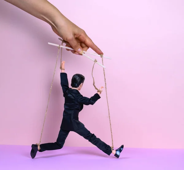 Pembe arka plan, manipülasyon ve kontrol kavramına sahip siyah takım elbiseli bir adamın kuklasıyla kadın eli.