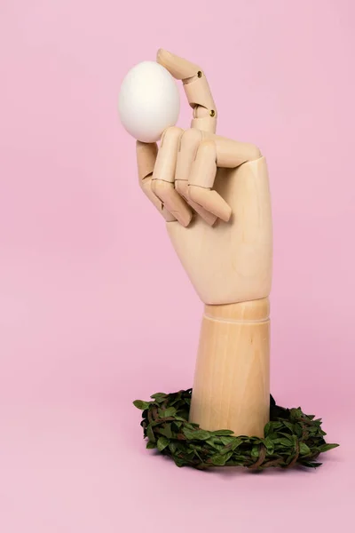 人类的木制手臂托着白色的蛋 装饰着春天的花环 彩绘粉红背景的复活节作文 — 图库照片