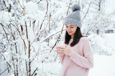 Gri şapkalı ve pembe kazaklı hoş, genç esmer kadın. Bir kış parkında elinde sıcak bir bardak içki tutuyor.