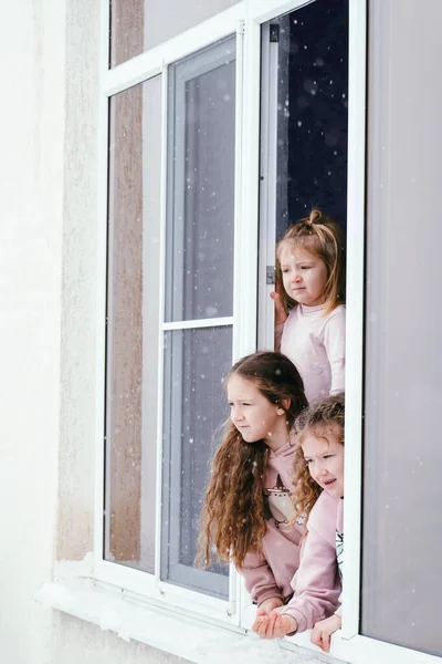 Three little girls lean out of the window and look at the falling snow