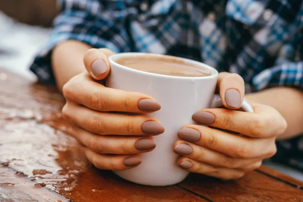 Close-up beautiful female hands hug white cup with hot drink on a wooden table outdoor.