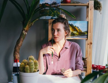 Genç ve güzel bir kadın çalışma masasında oturuyor ve yüzüne yakın bir fırça tutuyor. Fikirler ve ilham arayışı.