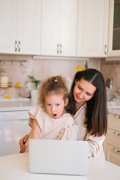 Cute portrait of mother and daughter sitting in a kitchen table and using computer.