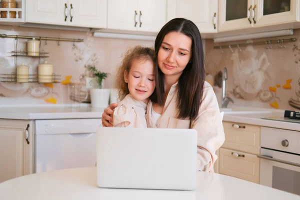 Cute portrait of mother and daughter sitting in a kitchen table and using computer.