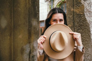Şirin, genç, esmer bir kadın, şapkasını yüzüne yakın tutuyor ve yürürken gülümsüyor.