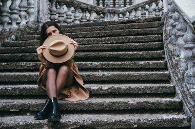 Bej şapka ve ceket giyen, merdivenlerde oturan güzel genç bir kadın..