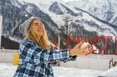 Mutlu genç kadın dişlek gülümseme, sarı saçlar, ekose gömlek ponpon şapkası kartopu oynuyor. Kış açık hava aktivitesi