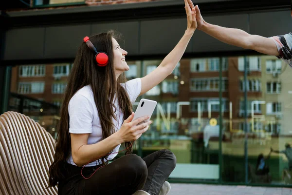 年轻美丽的女人用耳机和手机听音乐 在户外给她的朋友击掌 妇女坐在城市公园的长椅上 — 图库照片
