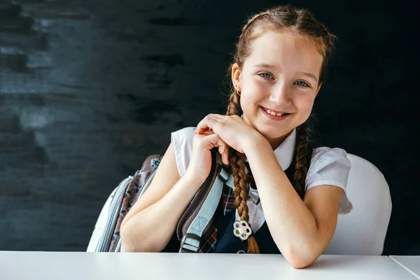 学校の制服を着たかわいい幸せな笑顔の若い女の子とバックパックは 空白の黒板の背景にテーブルの上に座っている 学校のコンセプトに戻る — ストック写真