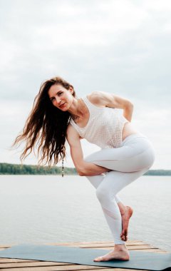 Beyaz spor elbiseli güzel bir kadın göl kenarındaki tahta bir piercingte egzersiz yapıyor..