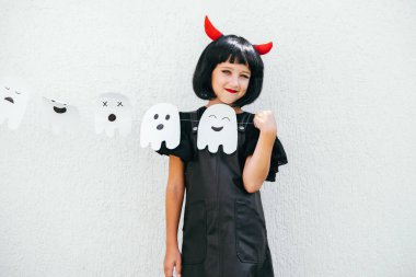 Dışarıda hayalet çelengi olan küçük bir kızın şirin Cadılar Bayramı portresi. Siyah elbiseli, kırmızı boynuzlu, siyah peruklu bir çocuk. Arkaplan için beyaz duvar.