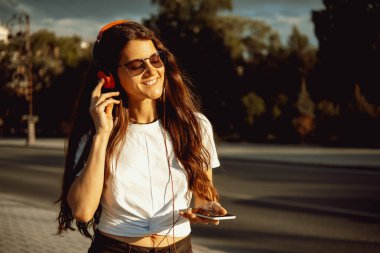 Güzel, mutlu, genç bir kadın cep telefonu, kulaklık kullanıyor ve şehir sokaklarında müzik dinliyor. Pozitif duygular, yaz aylarında hayat tarzı anlamlı portreler..