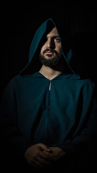 A man with a beard sits in a dark place looking at the camera and wears a blue traditional Moroccan djellaba.