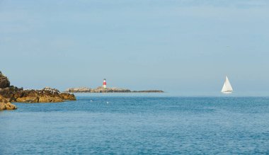 Old Muglins Lighthouse on the isolated island the backdrop of the blue cloudy sky, calm water and white sailboat.