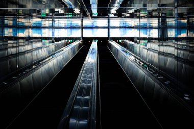 Ekranlardan parlak beyaz ve mavi ışık alan yürüyen merdivenlerin soyut karanlık sahnesi.