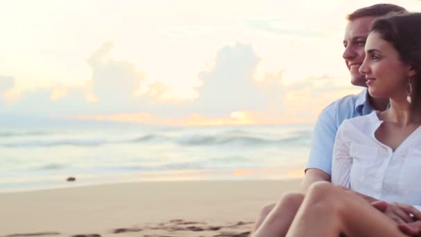 Hermosa joven pareja sentado juntos besándose en playa atardecer — Vídeo de stock