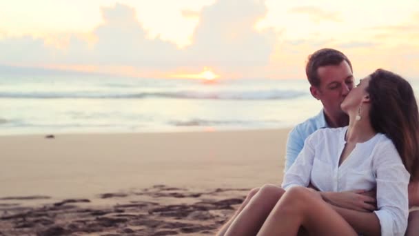 Hermosa joven pareja sentado juntos besándose en playa atardecer — Vídeo de stock