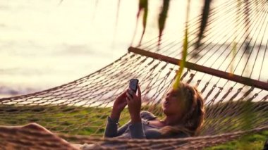 Young Woman Laughing Using Mobile Smart Phone while Relaxing in a Hammock by the Ocean Between Palm Trees at Sunset in Hawaii.