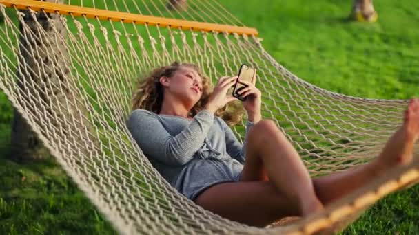 Modelo feminino atraente jovem com cabelo louro encaracolado usando telefone inteligente móvel enquanto relaxa em uma rede — Vídeo de Stock