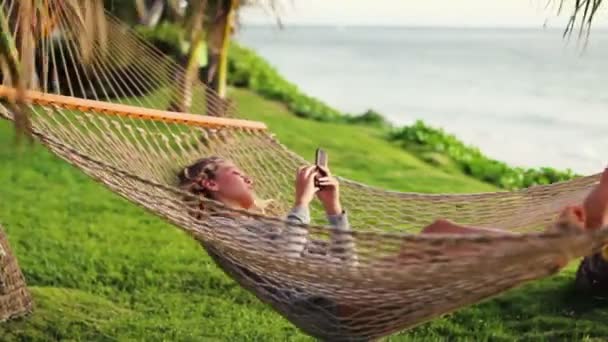 Onthullen pan van mooie blonde jonge vrouw met krullend haar met behulp van mobiele Smart Phone terwijl ontspannen in een hangmat — Stockvideo