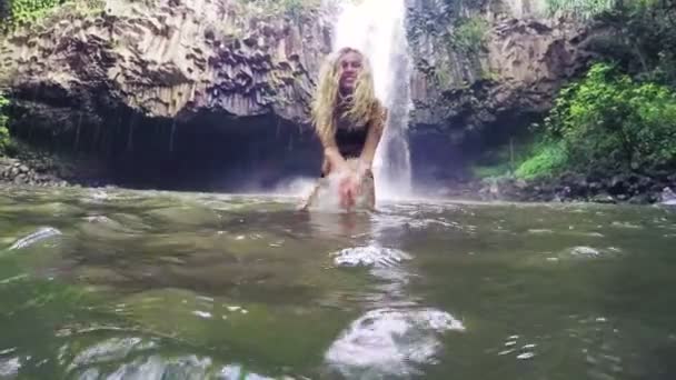 Hermosa joven modelo de bikini salpicando agua en la cámara en el exuberante estanque de la selva frente a la poderosa cascada — Vídeo de stock