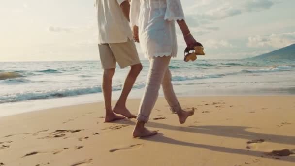 Sunset Walk em uma praia de luxo. Casal mais velho segura as mãos e caminha pela praia ao pôr do sol ficando com os pés molhados — Vídeo de Stock