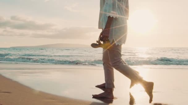 Закатная прогулка по роскошному пляжу. Пара пожилых людей держит руки и гуляет по пляжу на закате, моча ноги — стоковое видео