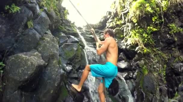 Felsen erklimmen Wasserfall. Junger Mann klettert im üppigen hawaiianischen Dschungel auf Seil — Stockvideo