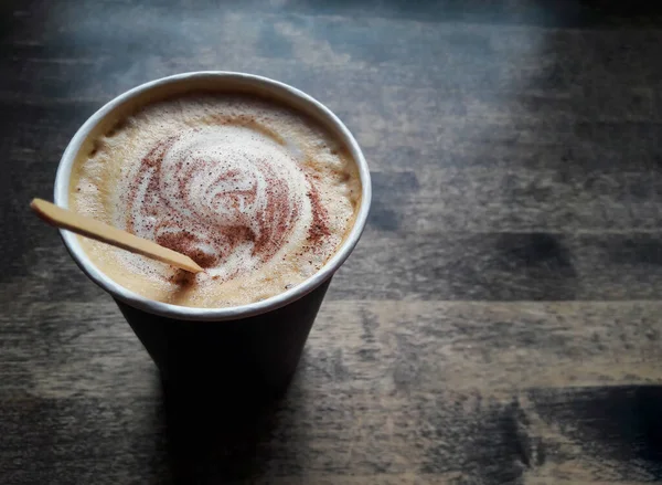 Cappuccino coffee with cinnamon on froth mixed wooden stick in a black paper cup stands on a wooden table near the window with natural light. Cappuccino closeup photo, top view.