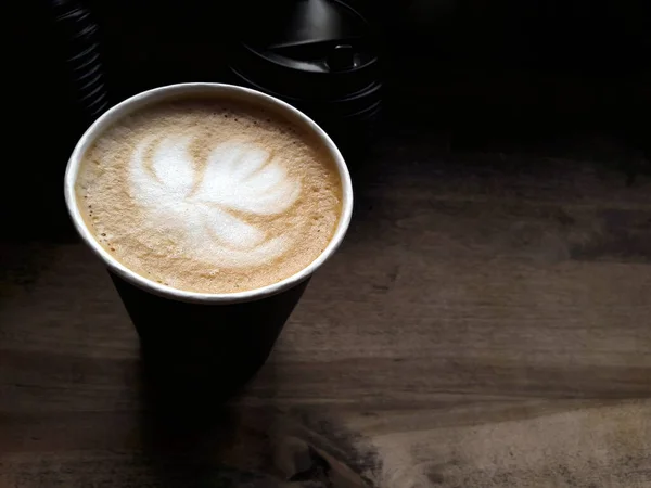 Siyah bir kağıt bardakta köpük üzerine nilüfer çiçeği çizilmiş bir Appuccino pencerenin yanındaki ahşap bir masada doğal ışık altında duruyor. Fotoğraf cappuccino yakın çekim, üst görünüm ve yan görünüm.