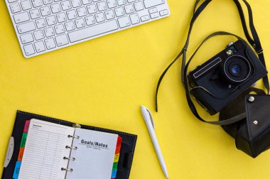 Çalışma alanı dairesi beyaz klavye, not defteri, not defteri, kalem ve klasik film kamerası sarı arka planda siyah deri çantada duruyordu. Karantina işyeri kavramı.