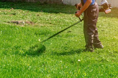 Koruyucu pantolonlu işçi çimenlikte çim biçme makinesiyle yeşil çim biçiyor..