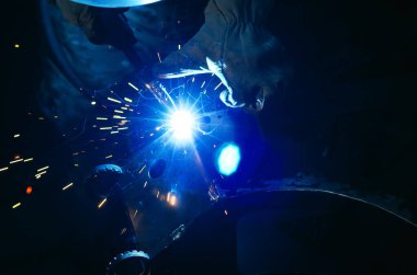 Parlak kıvılcımlarla kaynakçılığın mavi ışığı. Endüstriyel kaynak işlemi, kaynak işçisi koruyucu eldivenlerle karanlık arka planda. Metal kaynak fabrikası.