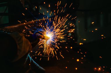 Sarı kaynak kıvılcımları etrafta uçuşurken işçi de koruyucu eldivenlerle metal parçalarını yapılandırıyor. Kaynak makinesi ile sürece yoğunlaşma ve hassasiyet.