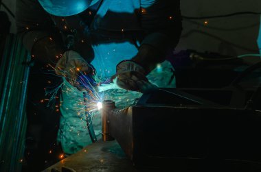Koruyucu giysili adam atölyede metal parçası kaynak yapıyor. Kötü aydınlatılmış endüstriyel odada kıvılcımlar, ışıklar ve duman. Kaynak.