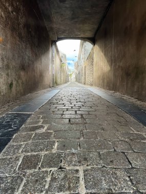 Dökümlü kaldırım taşlı dar sokaklar, eski yıpranmış duvarlar ve çıkışta yumuşak gün ışığı, derinlik, perspektif ve tarihi şehir atmosferi hissi yaratıyor..