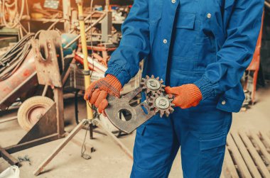 Mavi iş giysisi ve turuncu koruyucu eldiven giyen endüstriyel işçiler metal bir dişli mekanizması tutuyorlar. Üretim, makine mühendisliği, bakım kavramı.