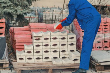 Mavi iş üniforması ve turuncu güvenlik eldiveni giyen bir kadın tahta paletlerin üzerine beton bloklar yığıyor. Endüstriyel malzemeler, el emeği ve inşaat alanı ortamı.