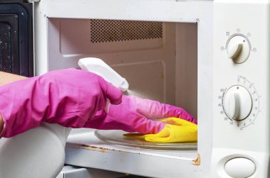 Koruyucu eldiven giyen bir kadın mikrodalga fırının içine temizlik solüsyonu sıkıyor. Ev hijyeni, bakteri temizliği, mutfak temizliği ve ev temizliği konsepti..