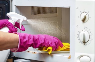 Koruyucu eldiven giyen bir kadın mikrodalga fırının içine temizlik solüsyonu sıkıyor. Ev hijyeni, bakteri temizliği, mutfak temizliği ve ev temizliği konsepti..