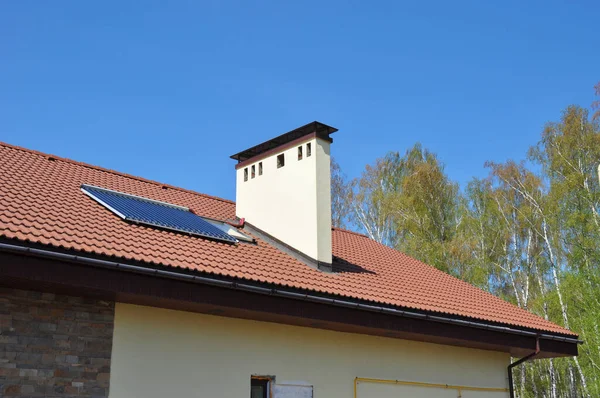 A close-up of an energy efficient detached house\'s clay roof with a solar water heating thermal panel, a skylight, a chimney and a rain gutter system against blue sky.