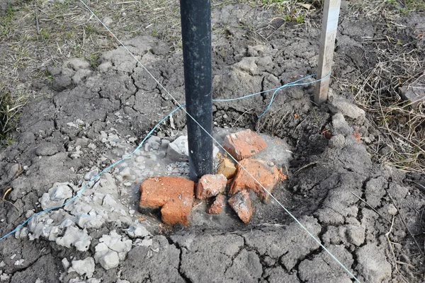 A close-up on a fence metal post installation, setting in concrete using a level line or string, crashed gravel, broken bricks and concrete to fill the post hole.