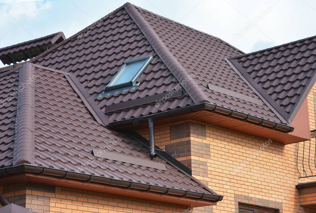 Complex roof design, metal tiled expensive roofing construction with a skylight, snow guards, chimney of a brick house.