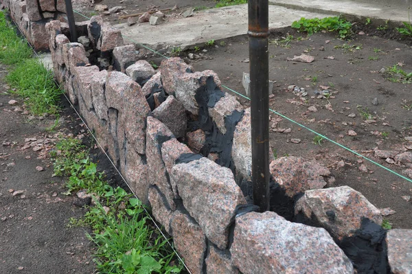 Brick fence construction and stone masonry: A close-up on the construction of a stone fence foundation, natural stone masonry footing along metal fence posts, and a level string line.