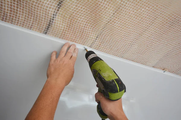 A builder is installing plastic ceiling panel, white PVC bathroom ceiling cladding in a bathroom using an electric screwdriver to attach it to the planked wood ceiling with a vapor barrier.